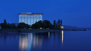 Een groot hotel met een modern design, gelegen aan een kalm waterlichaam dat het gebouw en de bomen weerspiegelt.