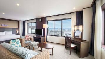 Habitación de hotel con cama, televisor, escritorio, silla, sofá y un amplio ventanal con vista a la ciudad.