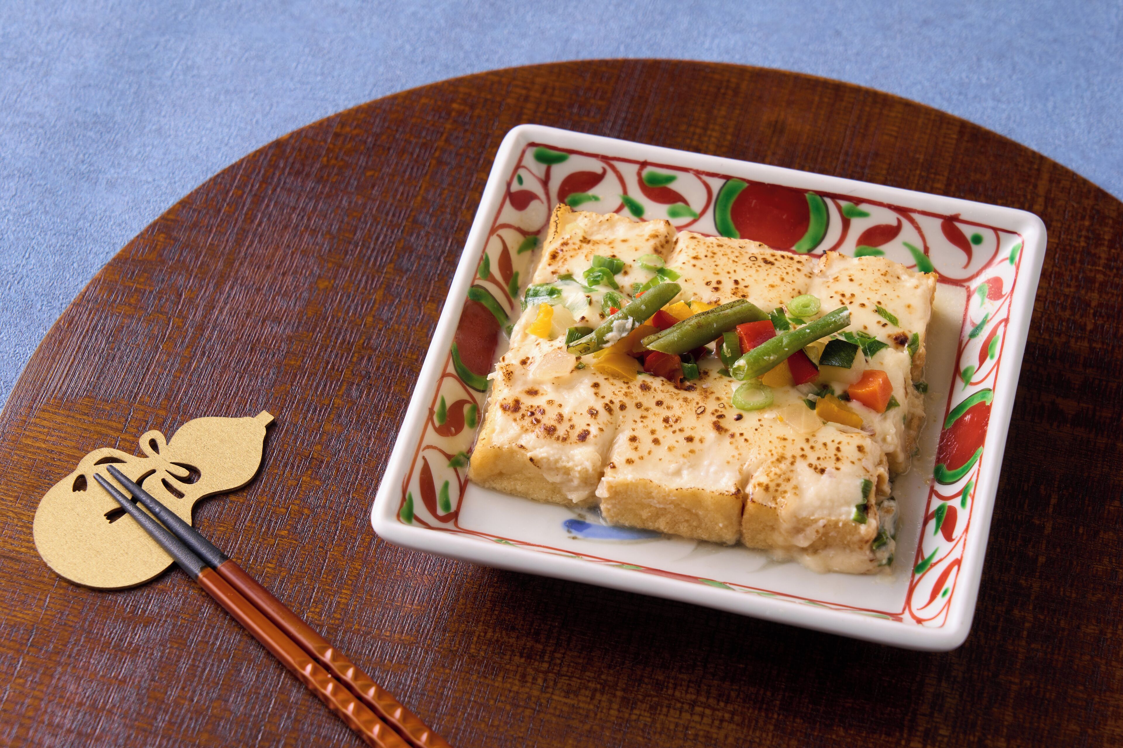 A square plate with a decorative border, containing a dish with tofu, green beans, and vegetables, accompanied by chopsticks and a decorative tag.