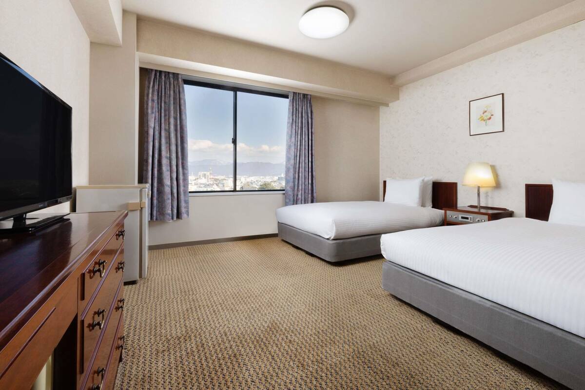 Habitación de hotel con televisión, dos camas, ventana con cortinas, lámpara y un cuadro en la pared.