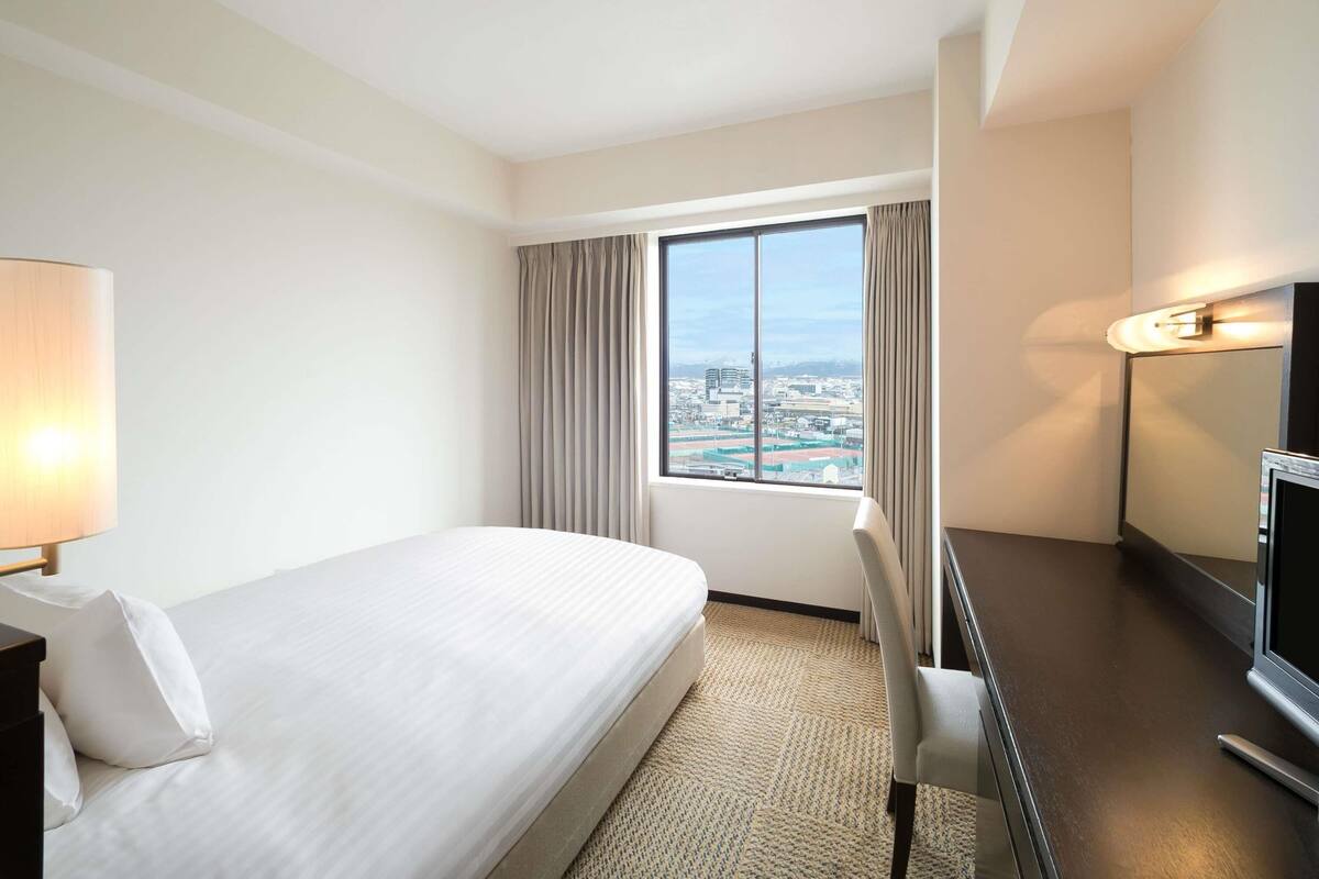 Habitación de hotel con cama, escritorio, silla, televisor y ventana con cortinas.