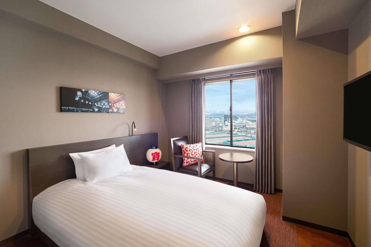 Habitación de hotel con una cama grande, una mesita, una silla, un televisor, un ventanal con vistas a la ciudad y un cuadro en la pared.