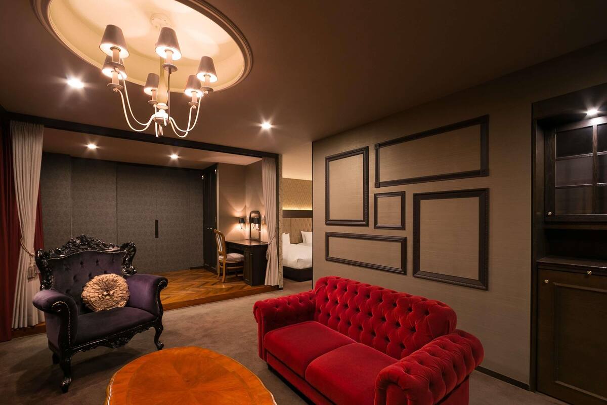 Una habitación con un sofá rojo, una mesa de centro de madera, un sillón negro, una araña y una cama con cabecera.
