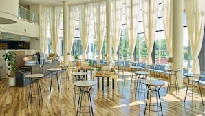 Un amplio espacio interior con techos altos, ventanas grandes con cortinas translúcidas, múltiples zonas de estar con mesas y sillas, y una zona de cocina con equipo de bar.