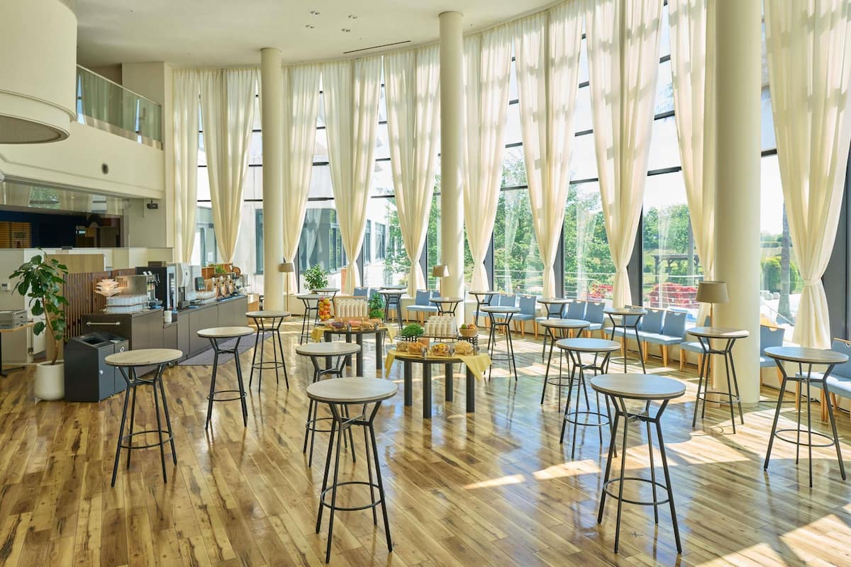 A spacious indoor area with high ceilings, large windows with sheer curtains, multiple seating areas with tables and chairs, and a kitchen area with bar equipment.