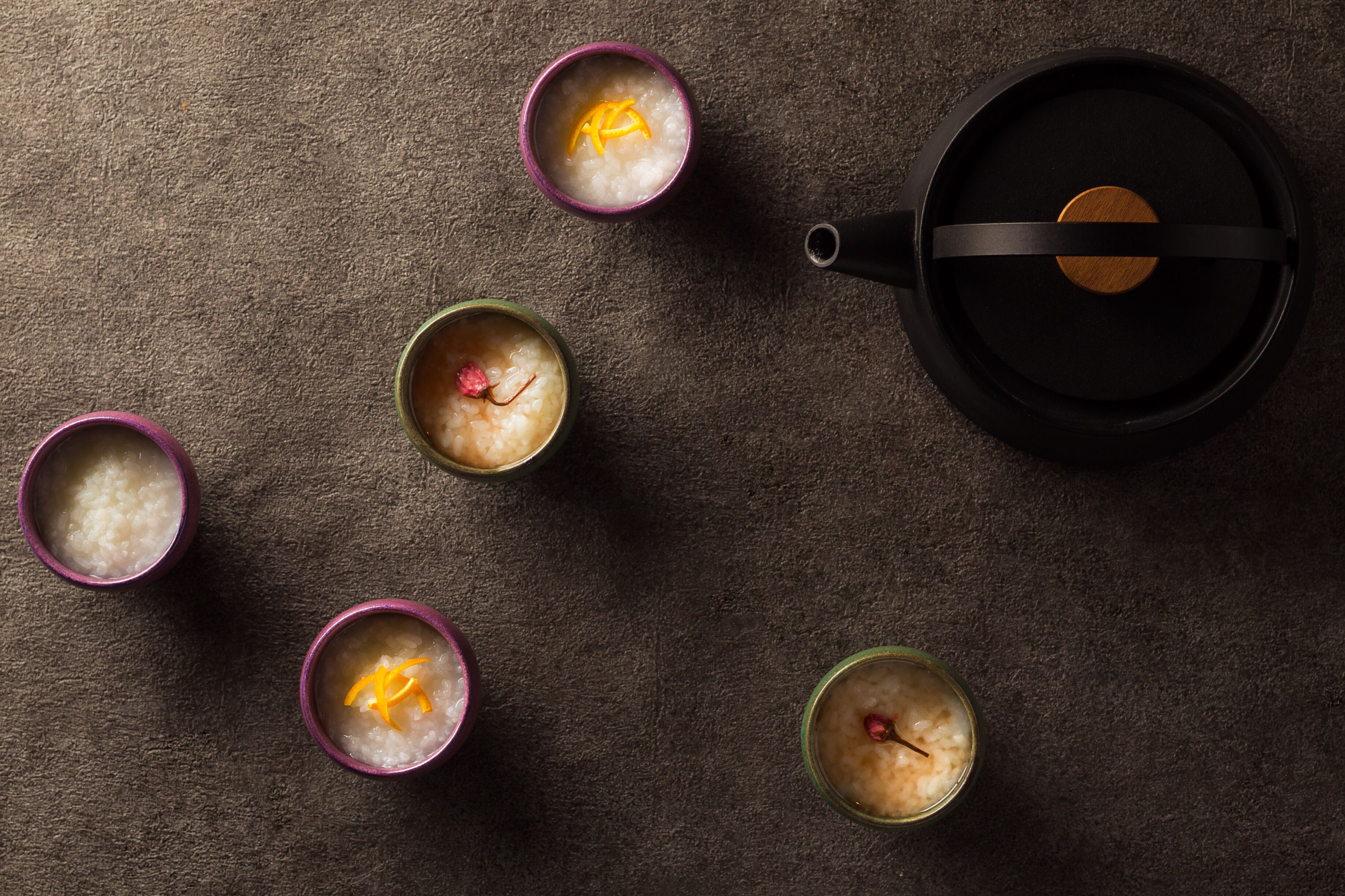 A top-down view of a dining table with five bowls of rice, each with a piece of orange peel and a red item, possibly a flower or fruit. A black teapot is also visible on the table.