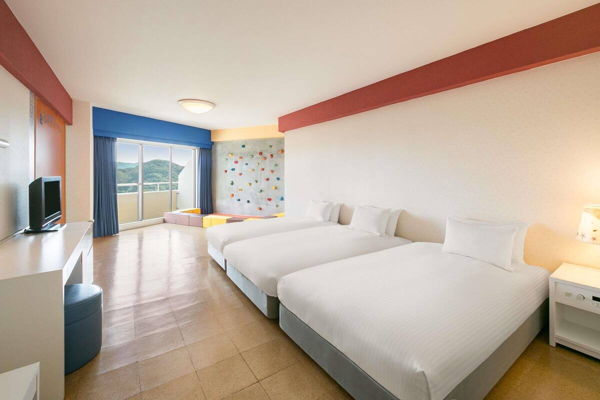 A hotel room with two beds, a TV, a desk, a large window, and a climbing wall.