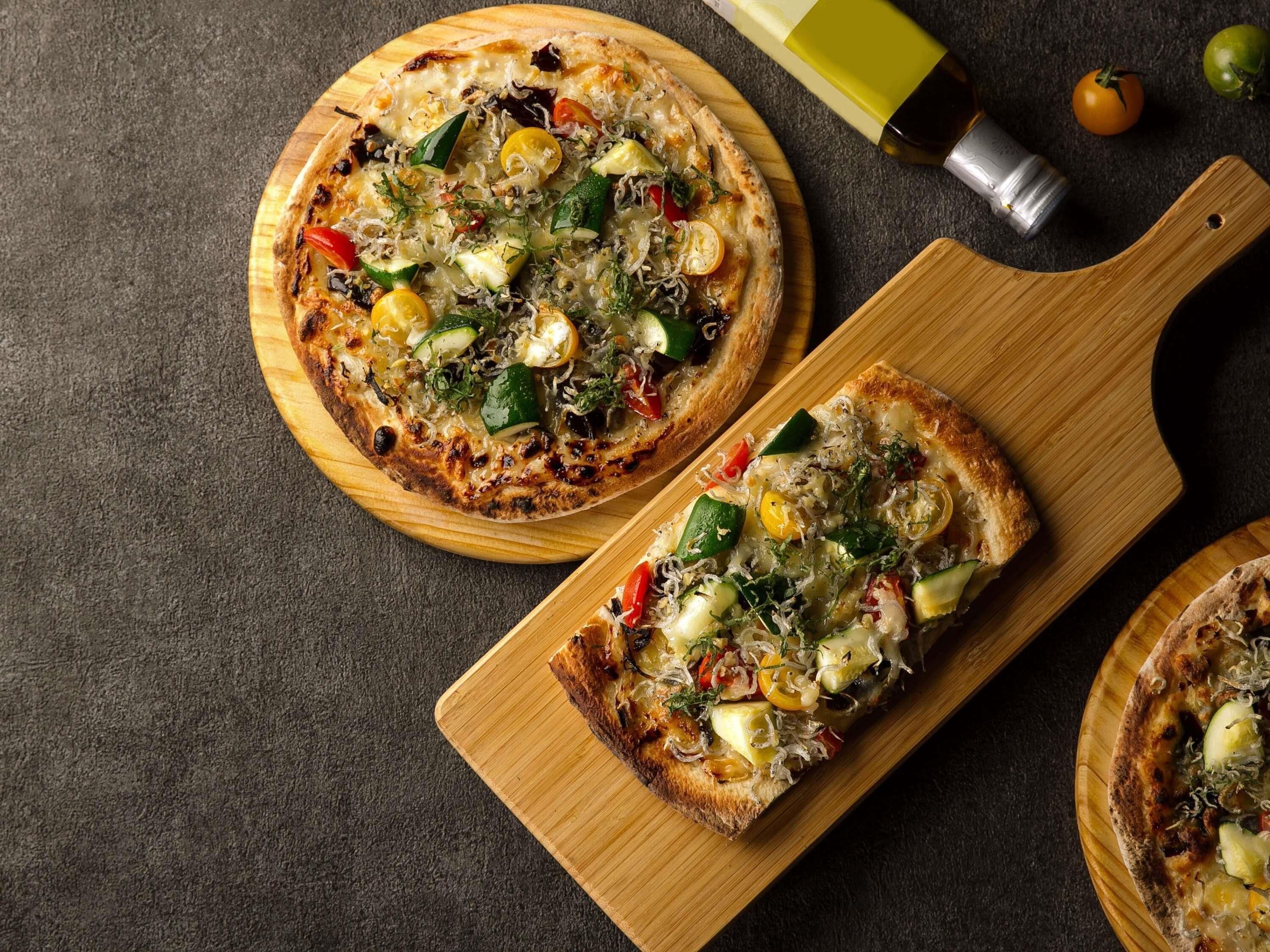 Three pizzas on wooden boards with a bottle and tomatoes in the background.