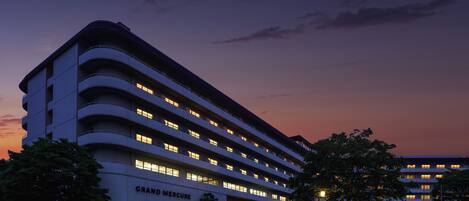 A multi-story hotel with a curved facade, named GRAND MERCURE, featuring a parking lot and several trees.