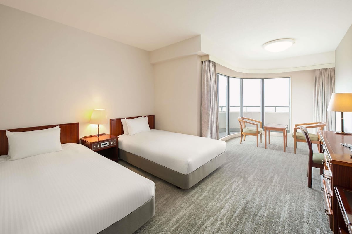 A hotel room with two beds, a desk, a chair, and a large window offering a view.