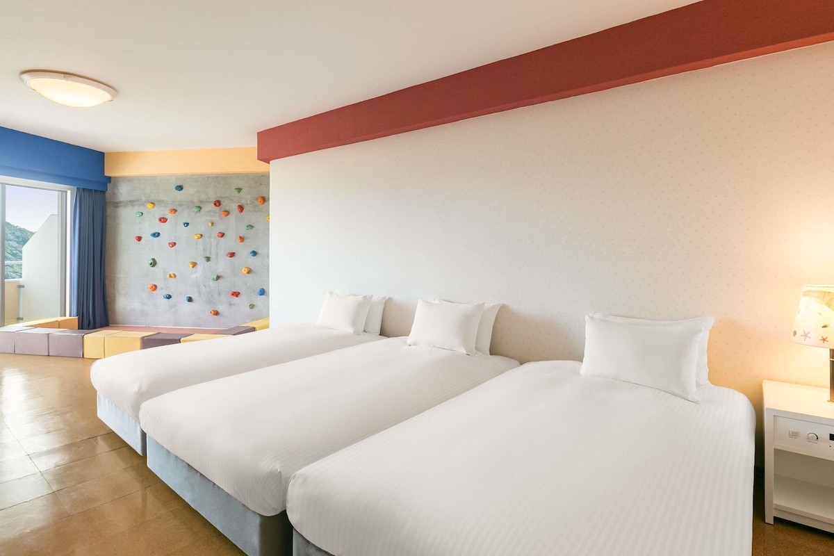 Habitación de hotel con dos camas, una pared de escalada, una ventana con cortinas y una mesita de noche con lámpara.