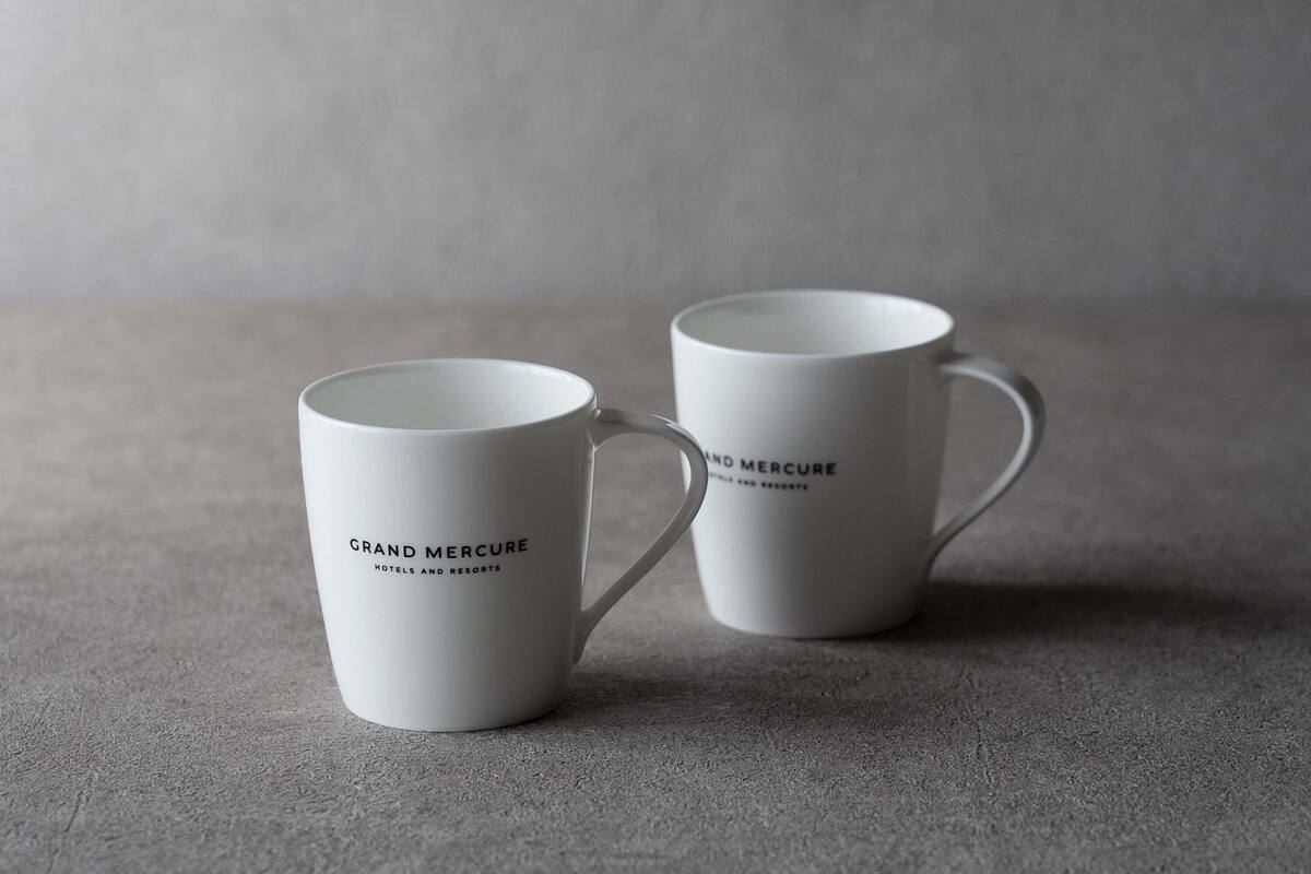 Two white mugs with the text "Grand Mercure Hotels and Resorts" on them, placed on a textured surface.