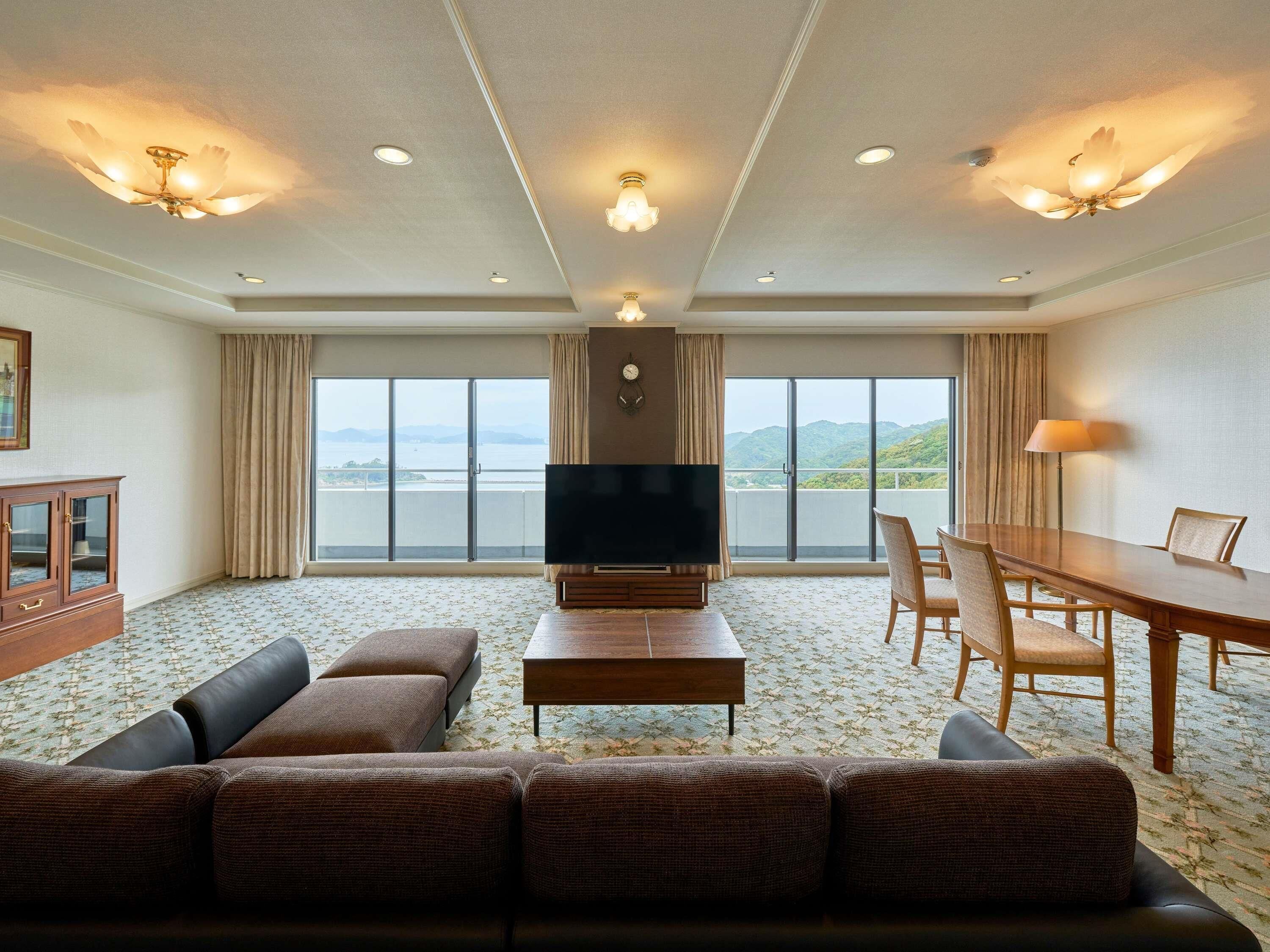 A hotel room with a large TV, a sofa, a dining table with chairs, and a view of the sea.