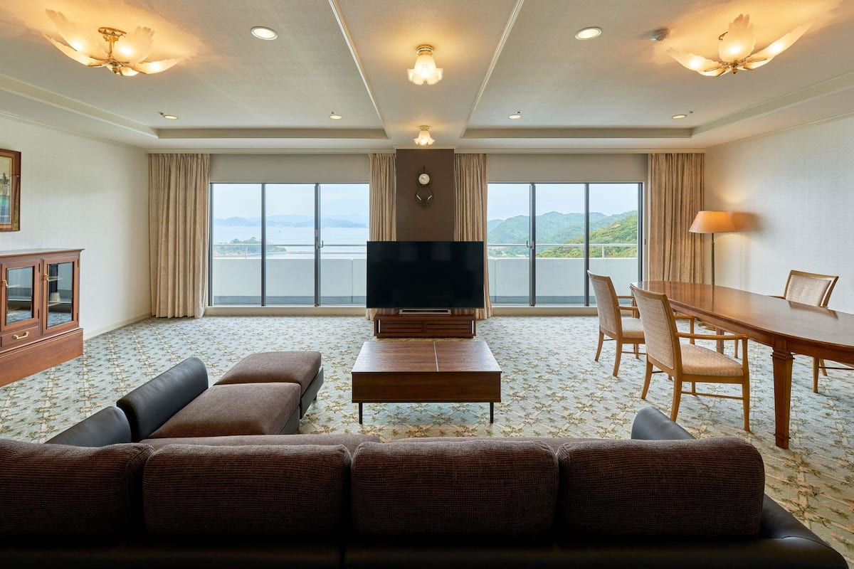 A hotel room with a large TV, a sofa, a dining table with chairs, and a view of the sea.