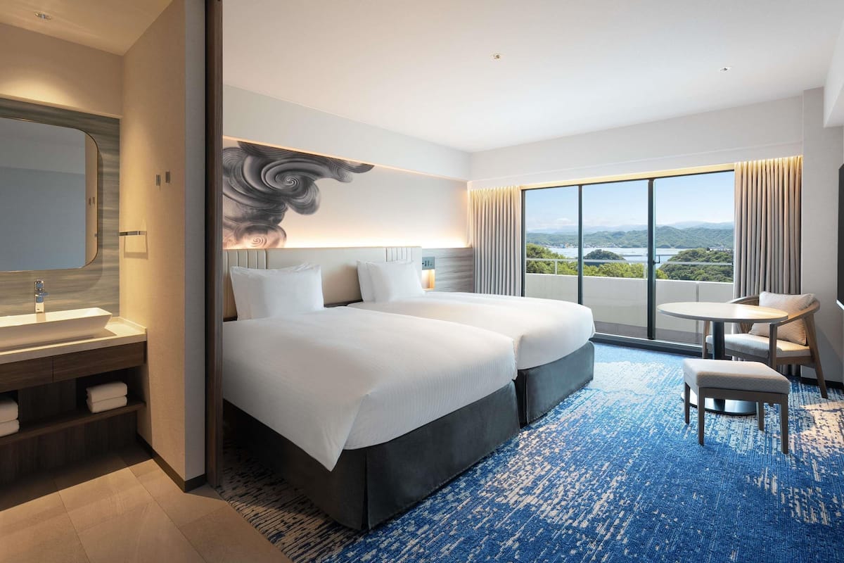 A hotel room with a large bed, a desk, a chair, a view of the outdoors, and a bathroom with a mirror and sink.