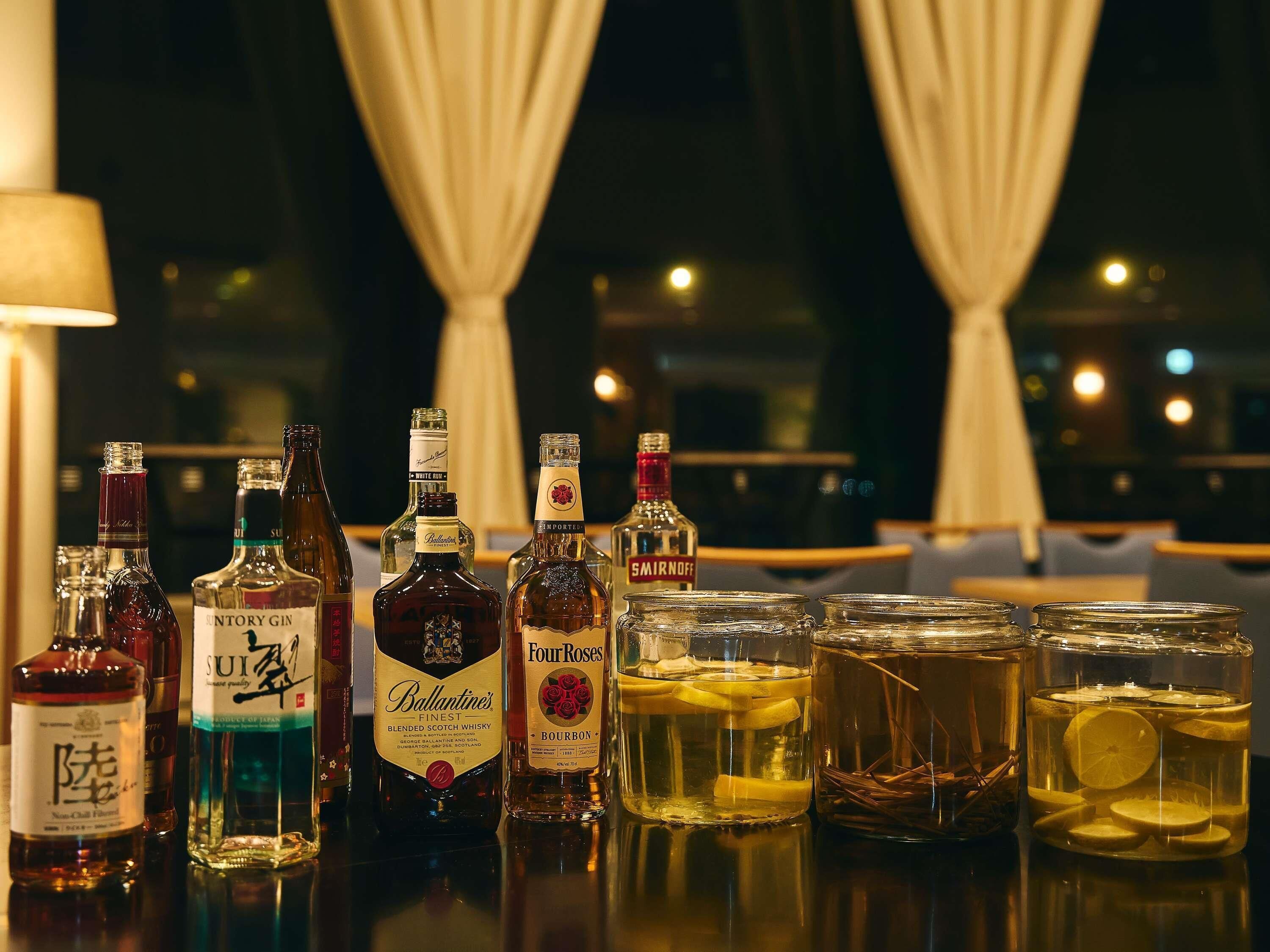 Variedad de bebidas alcohólicas y copas sobre una barra con vistas a cortinas y luces de fondo.