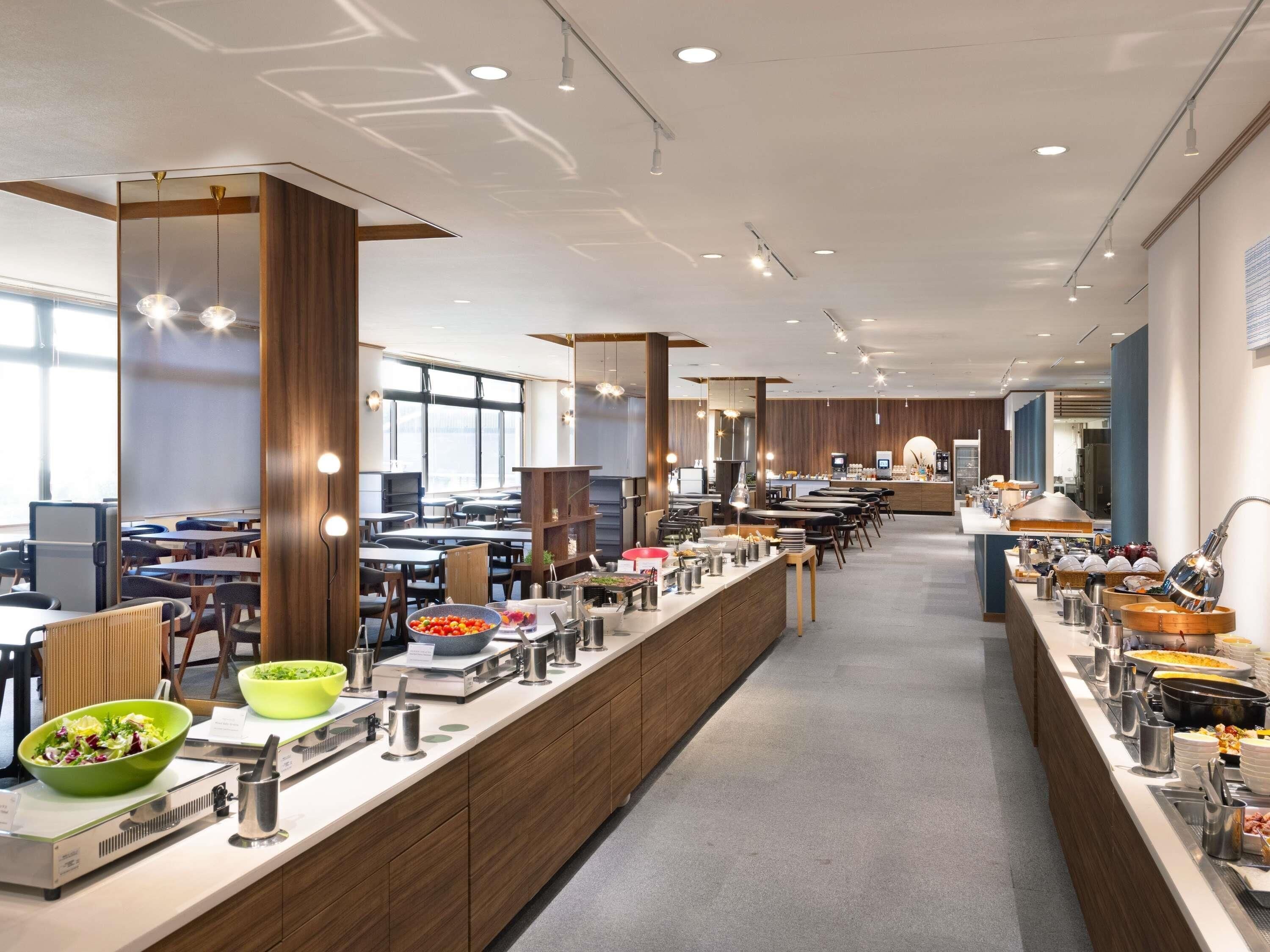 A modern cafeteria with long communal tables, a buffet line with various food items, and a seating area with individual tables and chairs.