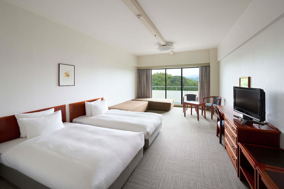 A hotel room with two beds, a desk with a TV, a chair, and a large window with a view of greenery.