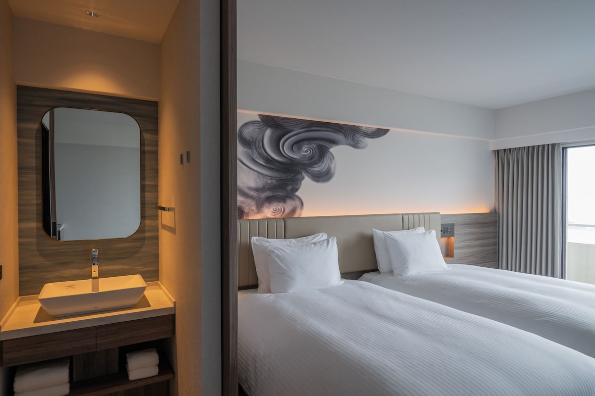 A modern hotel room with a large bed, a bedside table, a mirror, and a wall-mounted artwork.