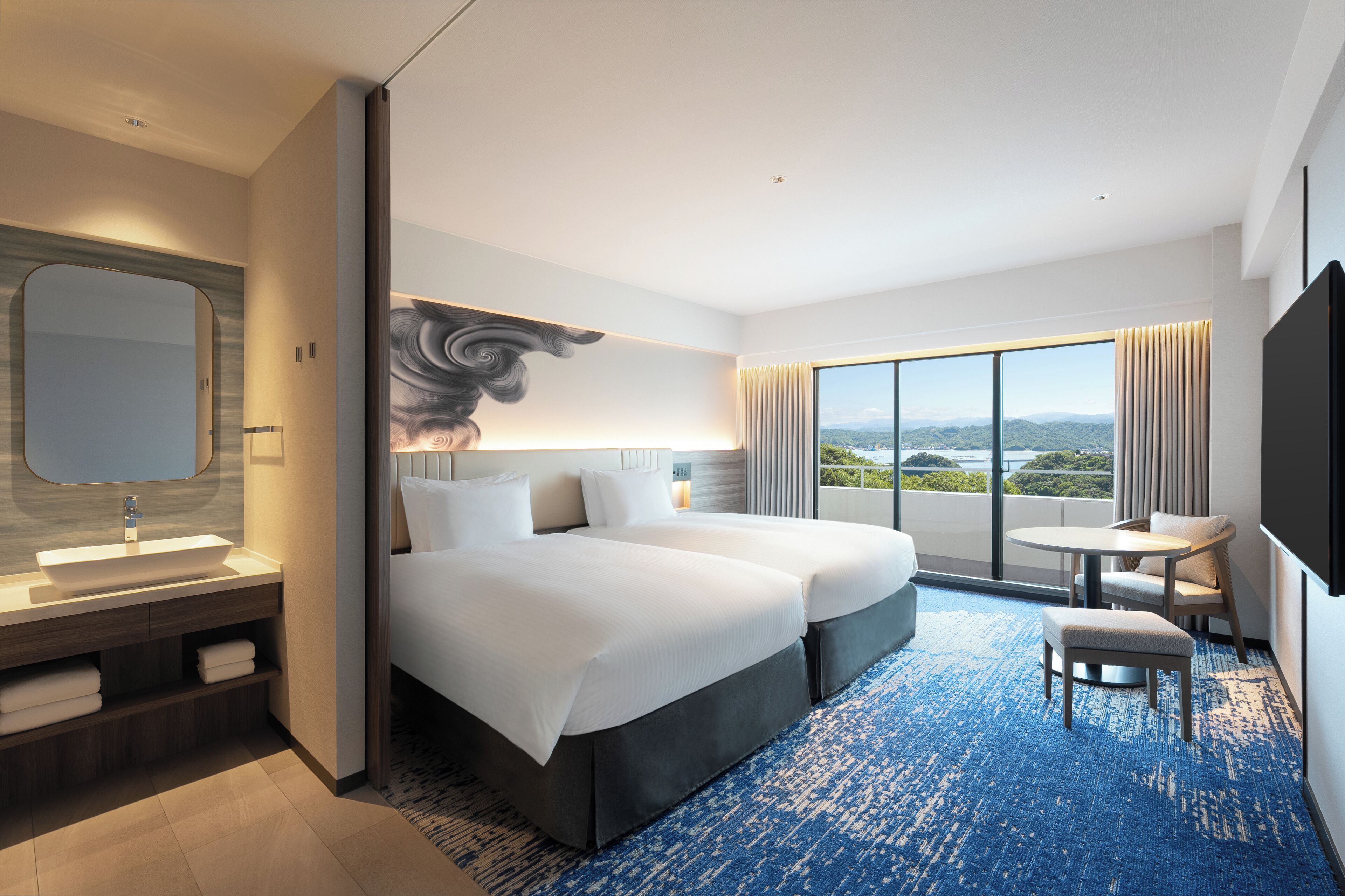 A modern hotel room with a large bed, a sitting area, a bathroom with a large mirror, and a view of the city.