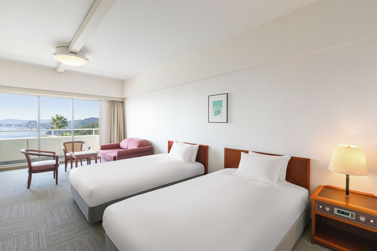 Habitación de hotel con dos camas, un escritorio, una silla, una mesita de noche con lámpara y un ventanal con vistas al exterior.