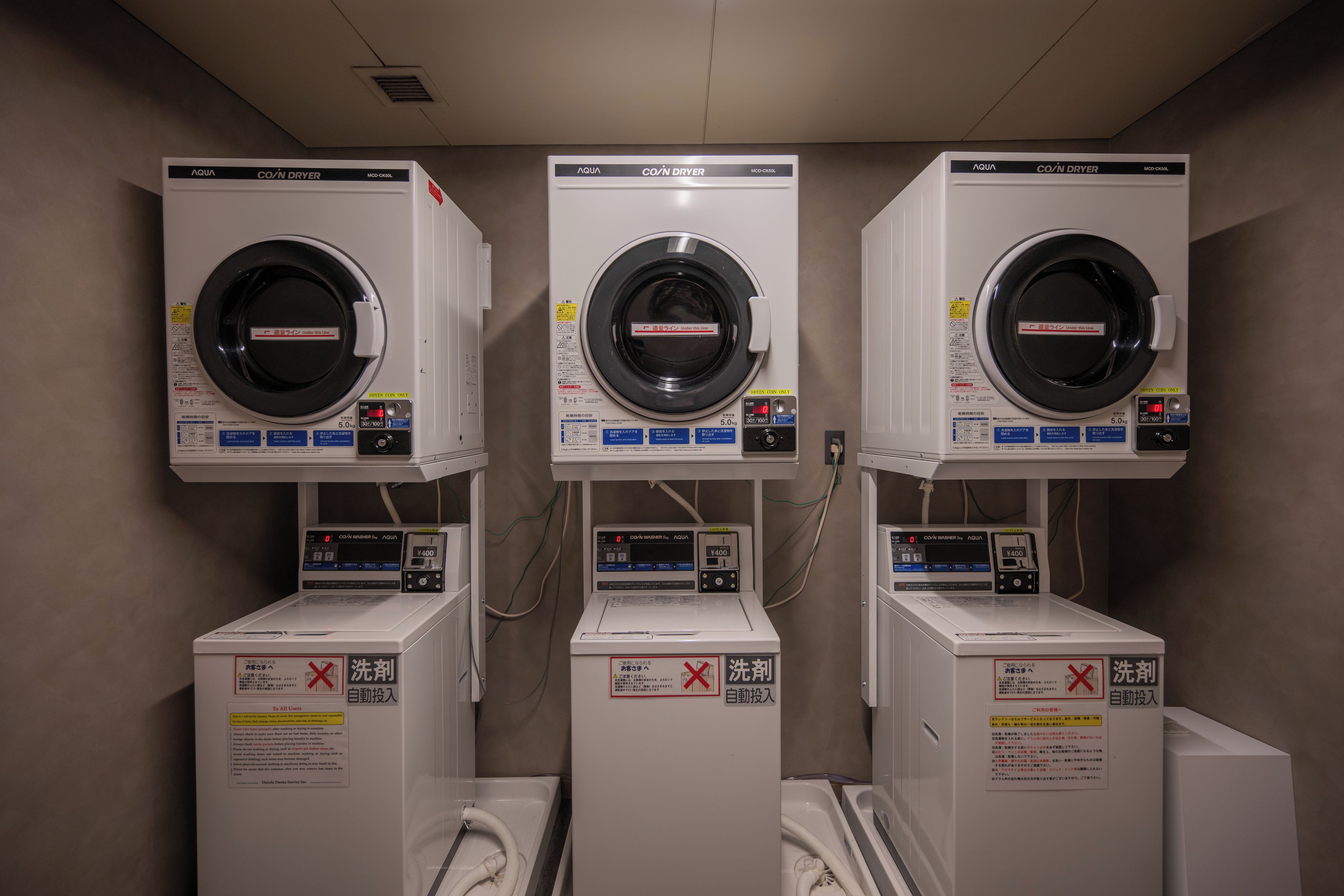 Three coin-operated washing machines in a row, each with a control panel and a warning sign.
