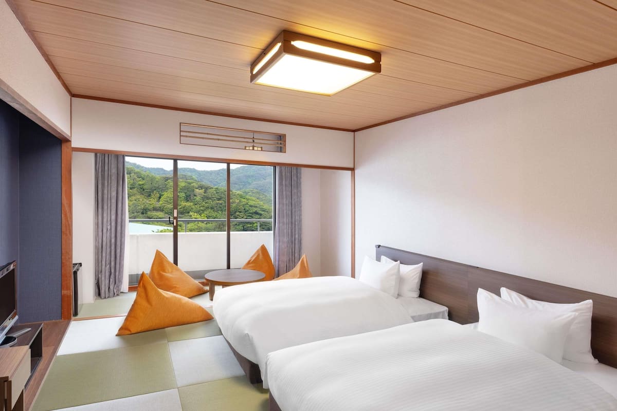 A hotel room with two beds, a small table, a TV, and a view of a mountainous landscape.