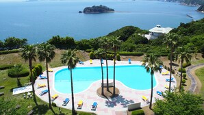 Un resort con una piscina grande rodeada de palmeras y sillas de descanso, con vista a una zona costera y montañas en la distancia.
