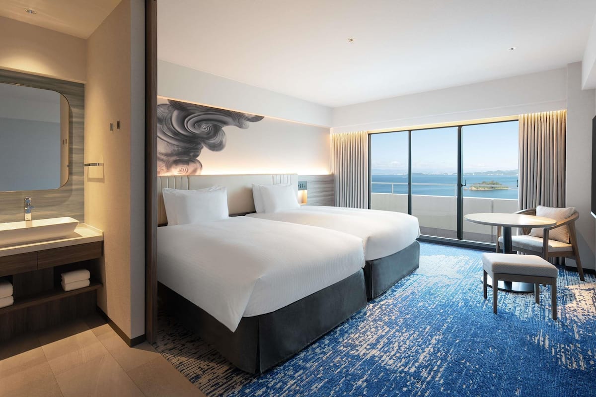 A hotel room with a large bed, a desk, a chair, a bathroom with a mirror, and a view of the sea.