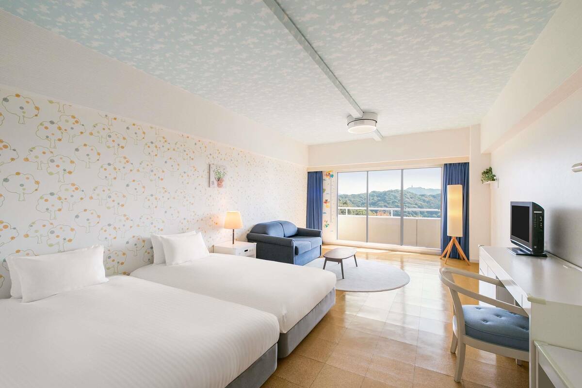 A hotel room with two beds, a desk, a chair, a lamp, and a large window with a view.