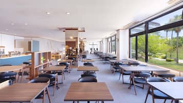 A modern dining area with tables and chairs, a kitchen area, and large windows offering a view of greenery.
