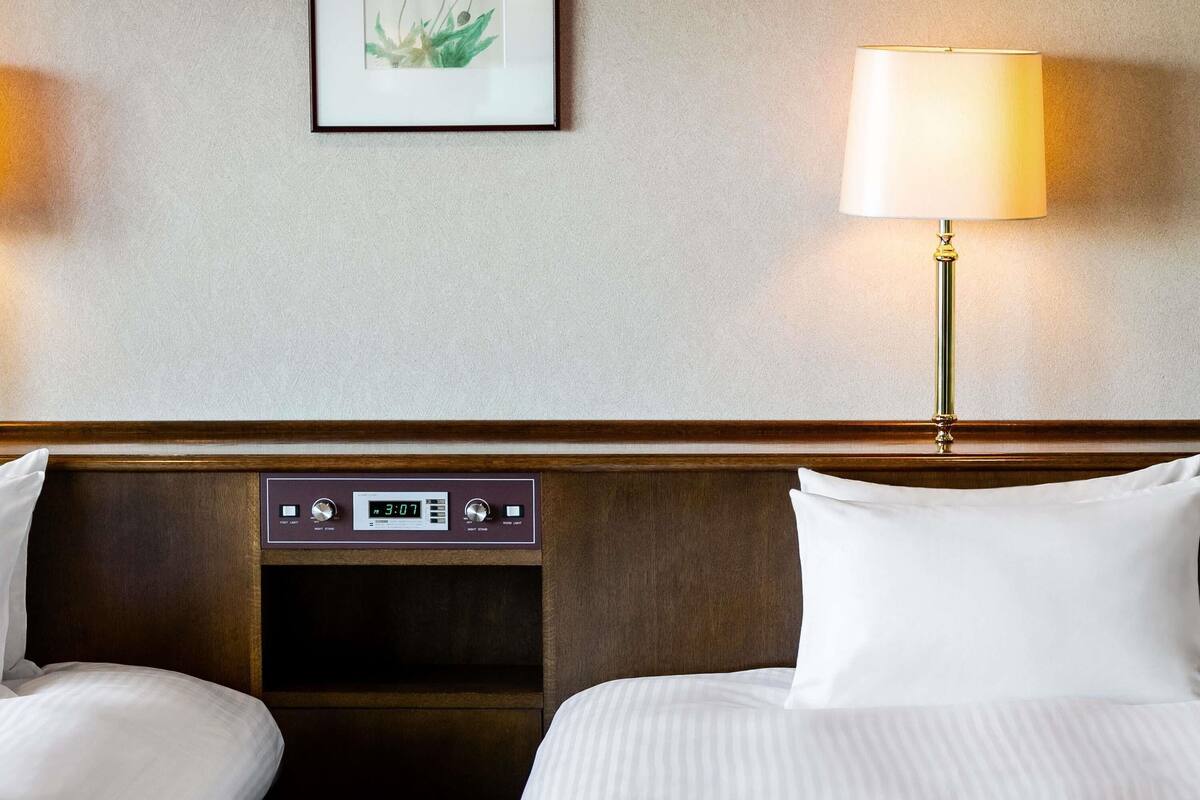 A hotel room with two beds, a bedside table with a clock, a lamp, and a framed picture on the wall.