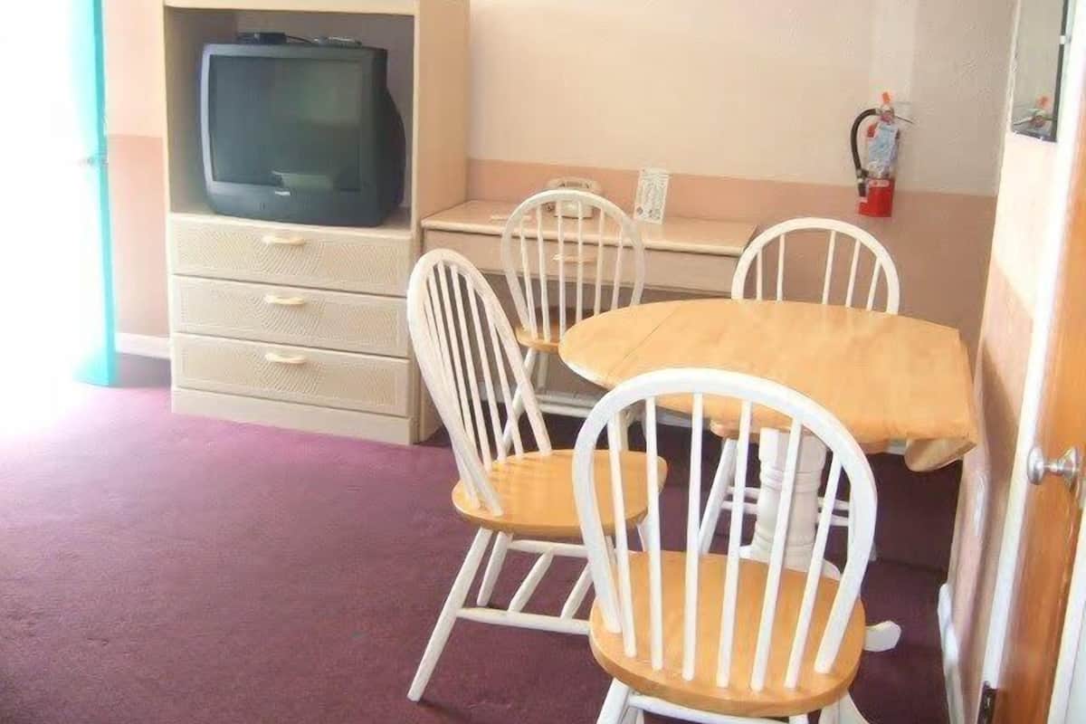 A room with a wooden table and four white chairs, a television on a cabinet, and a fire extinguisher on the wall.