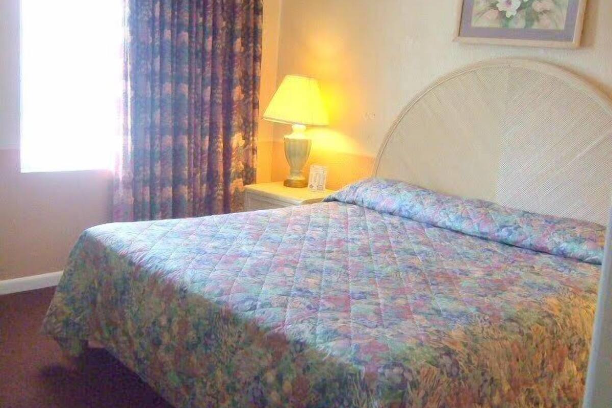 A hotel room with a bed, a nightstand, a lamp, a window with curtains, and a floral painting on the wall.