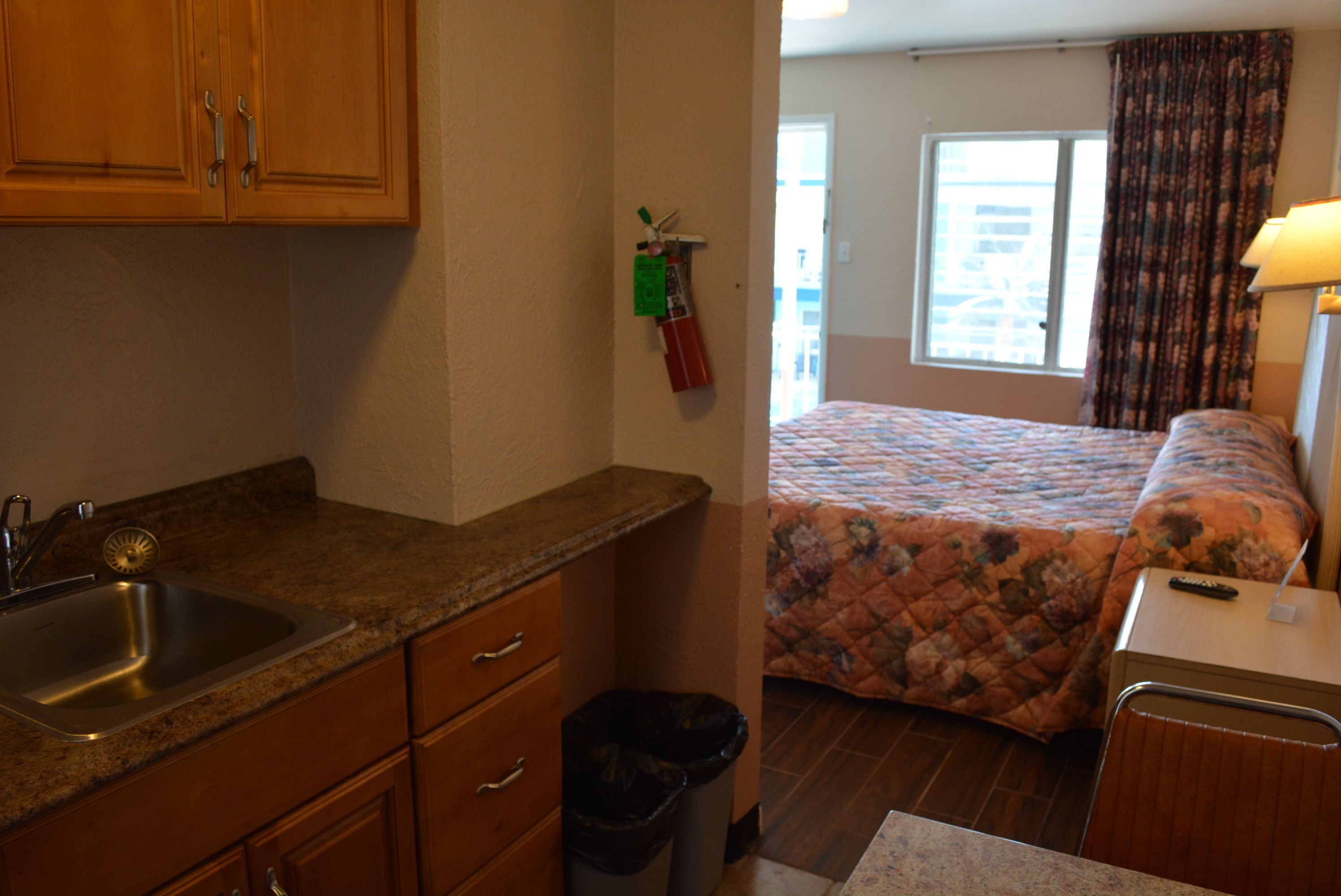 A compact hotel room with a kitchenette, a bed with a floral bedspread, a window with curtains, and a bedside table with a lamp.