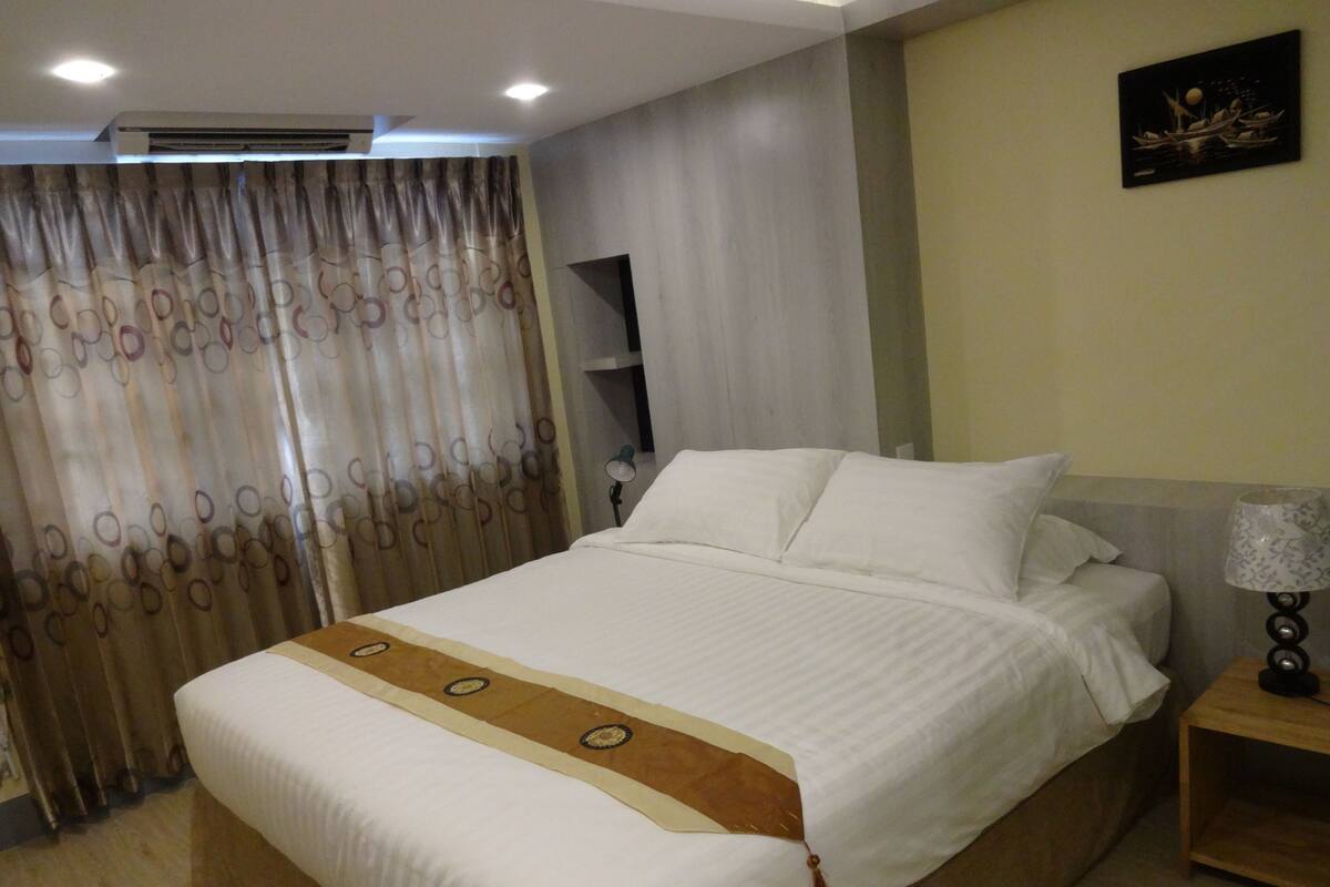 A neatly made bed with white linens and a brown decorative strip, a bedside table with a lamp, and a curtain with a circular pattern.
