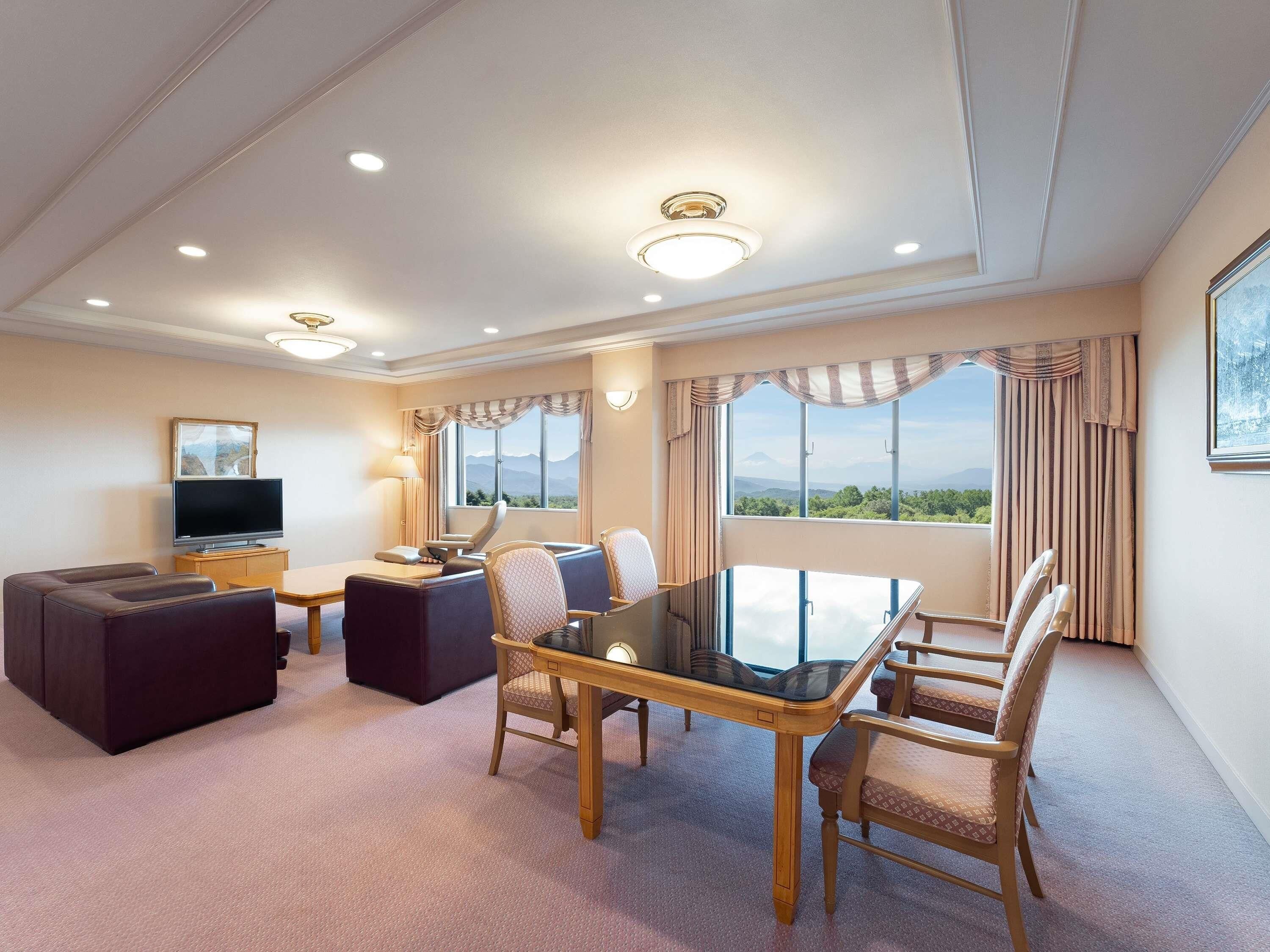 A hotel room with a dining table, chairs, a sofa, a TV, and a large window with a mountain view.