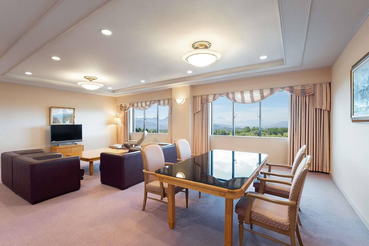 A hotel room with a dining table, chairs, a sofa, a TV, and a large window with a mountain view.