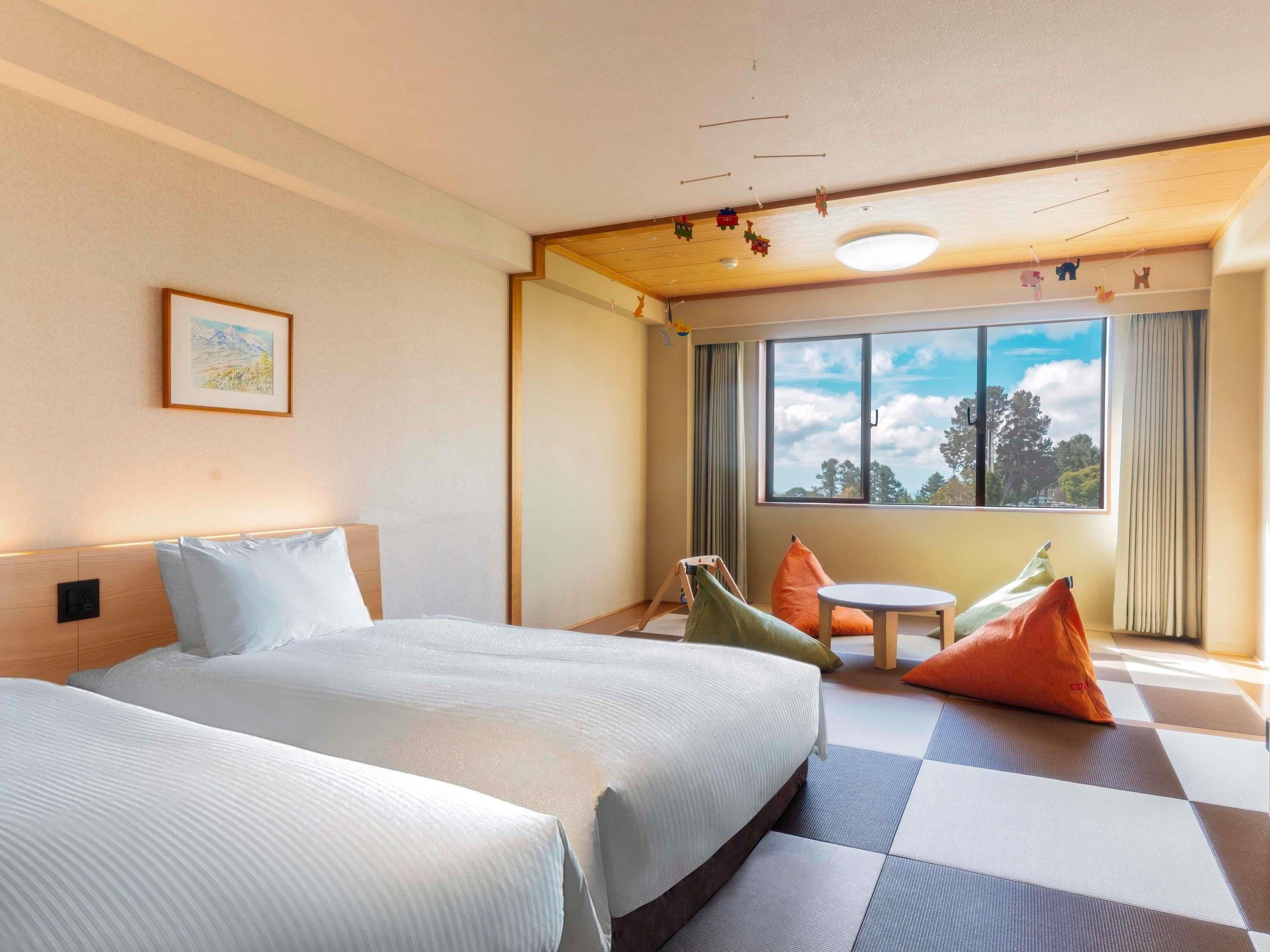 Habitación de hotel con dos camas, una mesita pequeña, cojines de colores, una ventana con vistas y una lámpara en el techo.