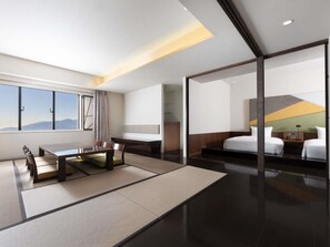 A modern hotel room with a dining area, a large window with a mountain view, a bed with a headboard, and a bedside table.
