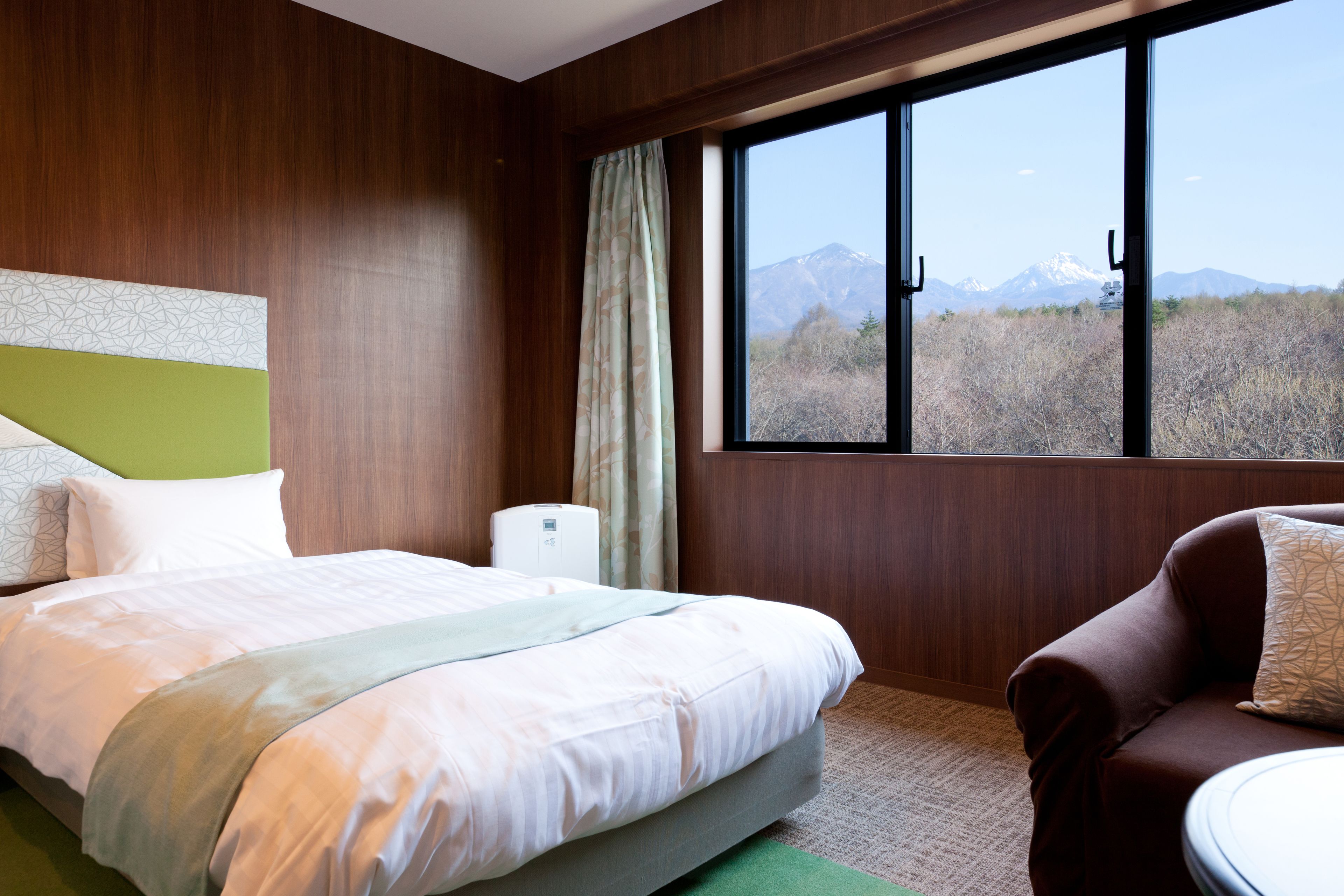 Habitación de hotel con una cama, cabecera verde, ventana con vistas a la montaña, sofá marrón y una mesita.