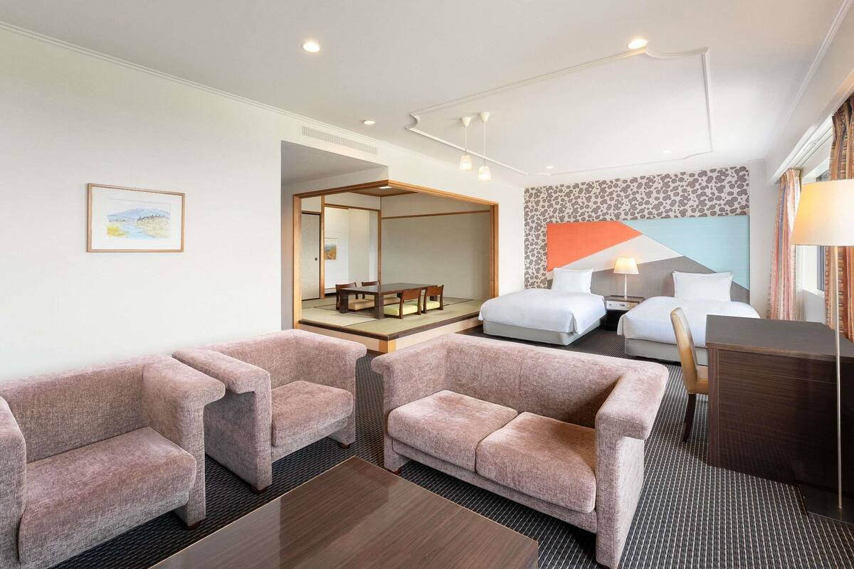 Habitación de hotel con una cama, dos sillones, una mesita, un escritorio, un espejo y un cuadro en la pared.