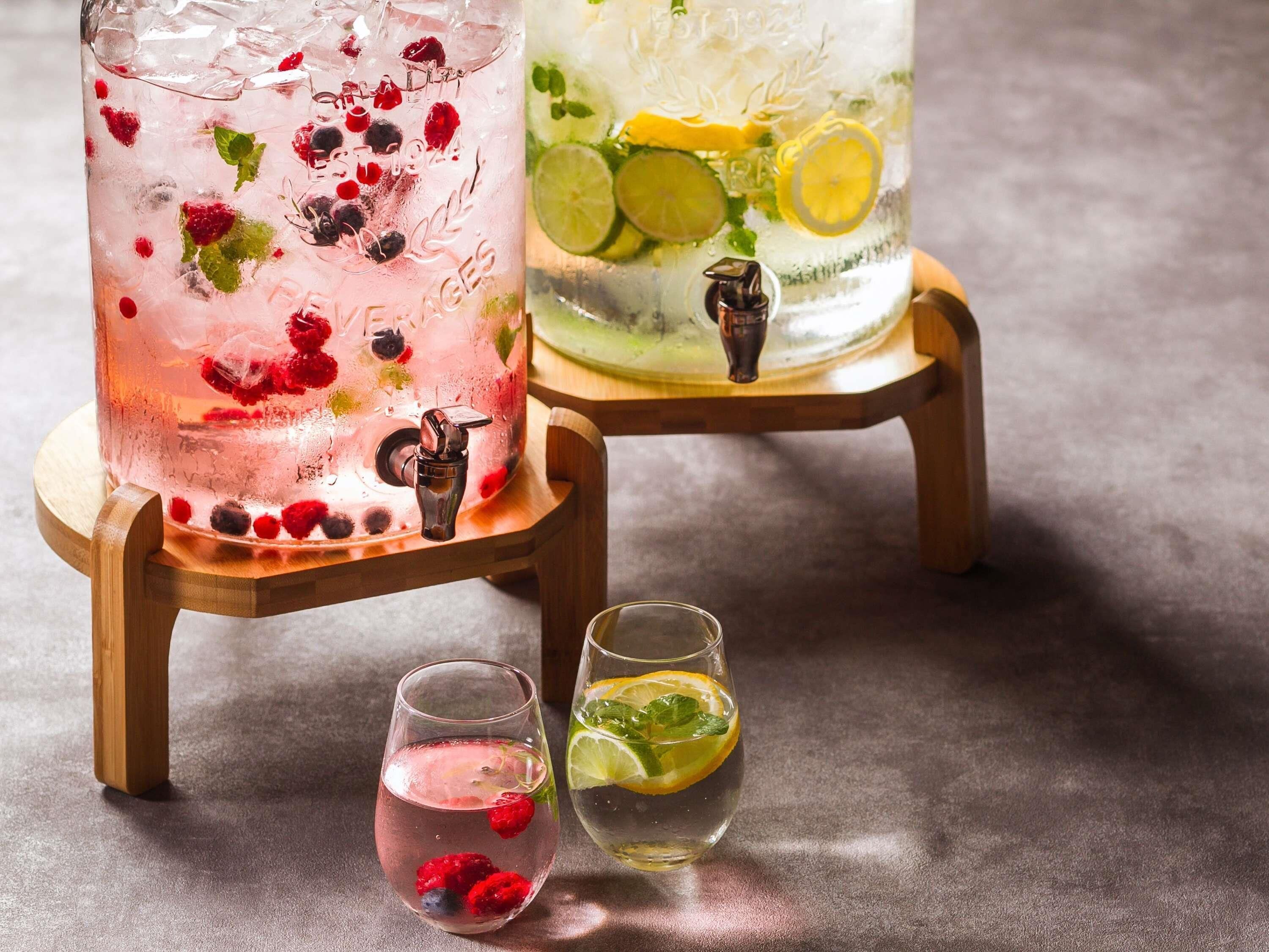 Two large glasses of iced beverages on wooden stands, one with berries and mint, and the other with lime slices and mint.