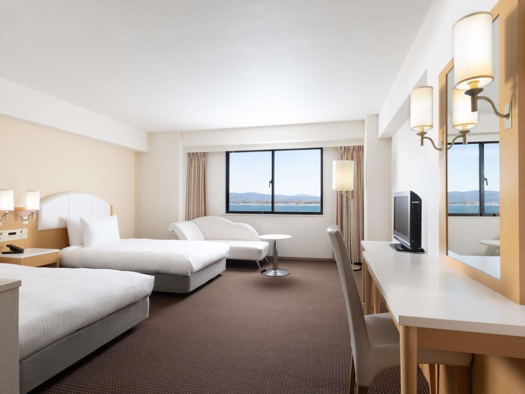 Habitación de hotel con dos camas, un escritorio, una silla, un televisor y un amplio ventanal con vistas.