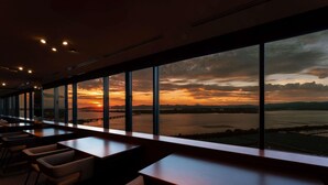 Restaurante com grandes janelas oferecendo vista para um pôr do sol sobre a água.