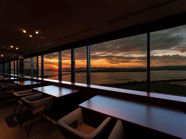 Un restaurante con amplios ventanales que ofrece vistas a un atardecer sobre el agua.