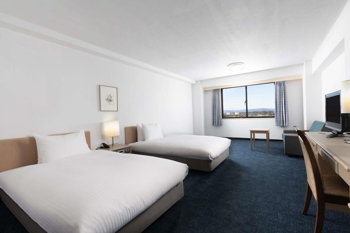 A hotel room with two beds, a desk, a chair, a lamp, a window with a view, and a picture on the wall.