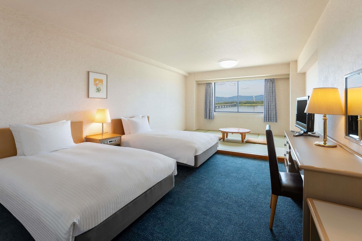 A hotel room with two beds, a desk, a chair, a TV, a lamp, a small table, and a window with a view of a bridge and mountains.
