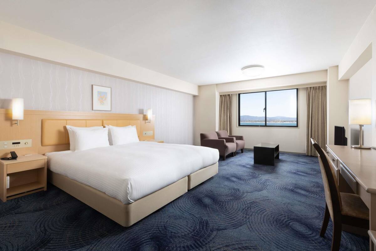 A hotel room with a large bed, a desk, a chair, and a view of the outdoors.
