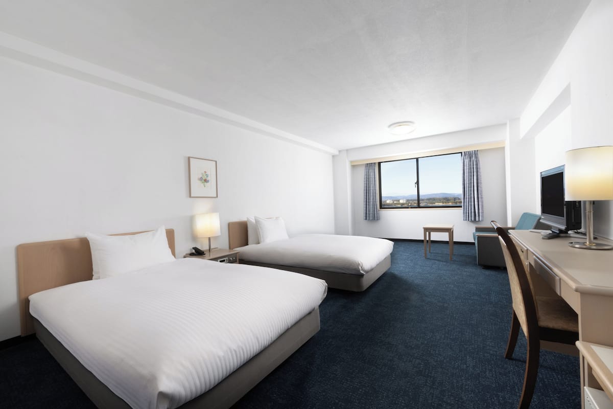 A hotel room with two beds, a desk, a chair, a TV, and a window with a view.